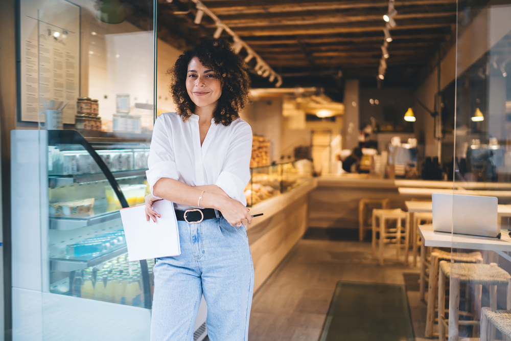 Woman business owner standing in cafe holding a paper with arms crossed, representing franchise for e2 business.