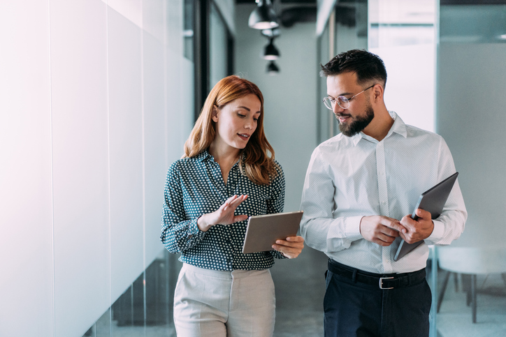 A man and woman are walking down an office hallway while discussing business strategy.