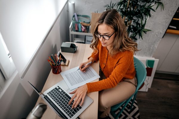 A woman with glasses types on a laptop while while looking at a handwritten copy of an O-1 recommendation letter.