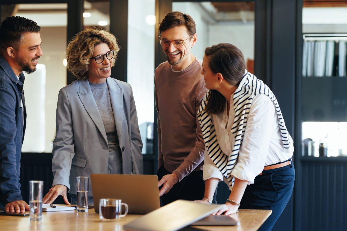 A group of executive business people stand around a table with a laptop and talk during a meeting.