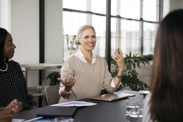 A mature professional woman gestures that she is weighing two options during a business meeting.