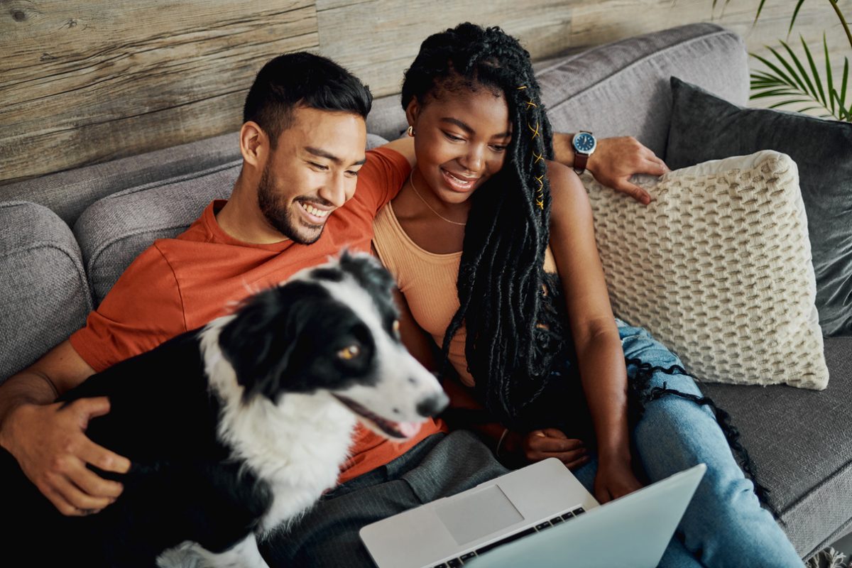 A married man and woman look at a laptop while sitting on a couch with their dog and smiling.