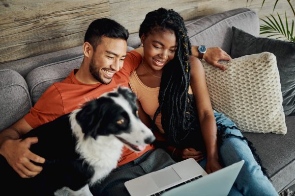 A married man and woman look at a laptop while sitting on a couch with their dog and smiling.