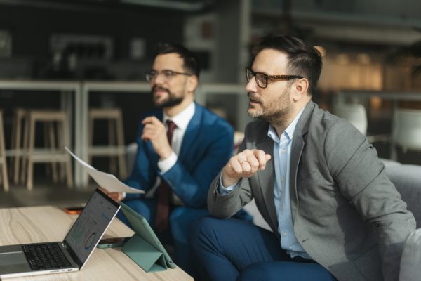 Two businessmen investors are discussing an investing strategy while looking at a laptop and documents.