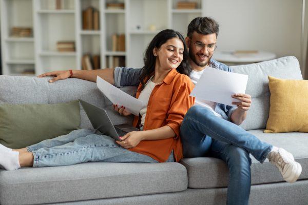 A married man and woman sit on a couch, smiling and looking at documents together.