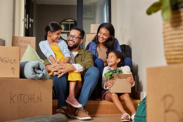 A young mother, father, and two children sit in the doorway of a house, surrounded by boxes after moving.