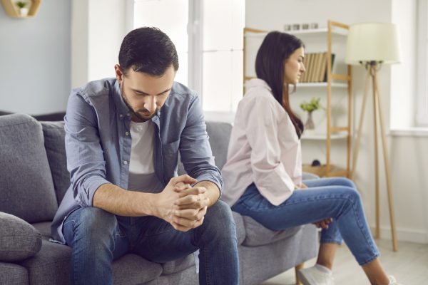 An upset married man and woman sit on a sofa looking away from each other.