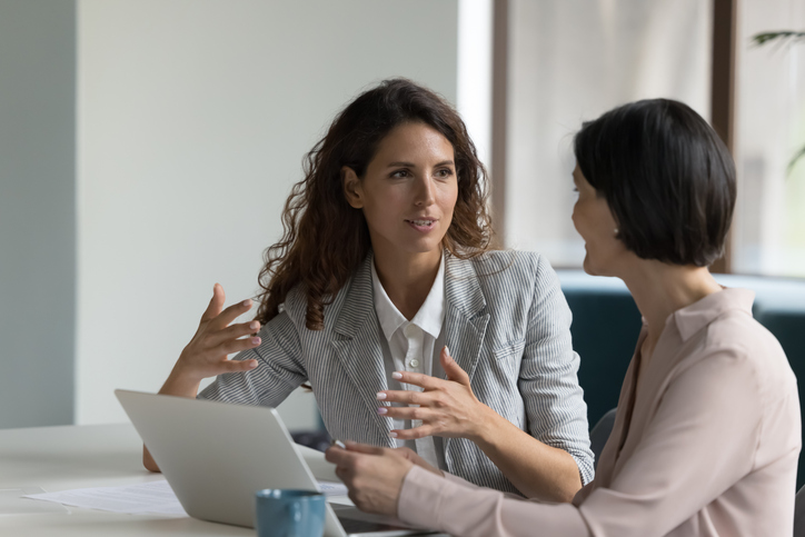 Two business women sit at desk discussing project details.