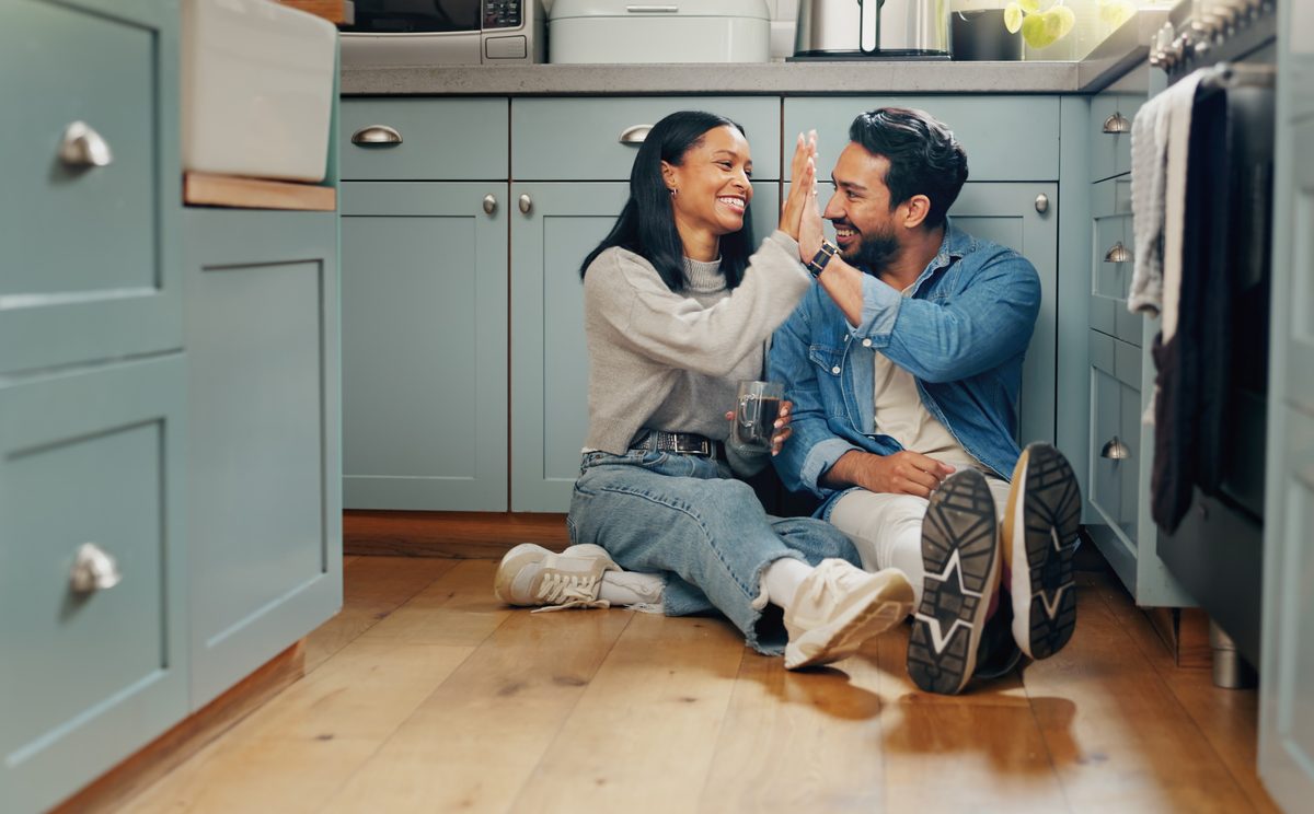 Happy married couple sitting on the floor of their kitchen clasping hands together.