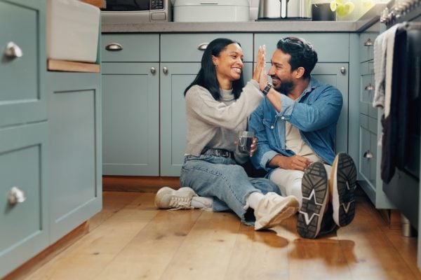 Happy married couple sitting on the floor of their kitchen clasping hands together.