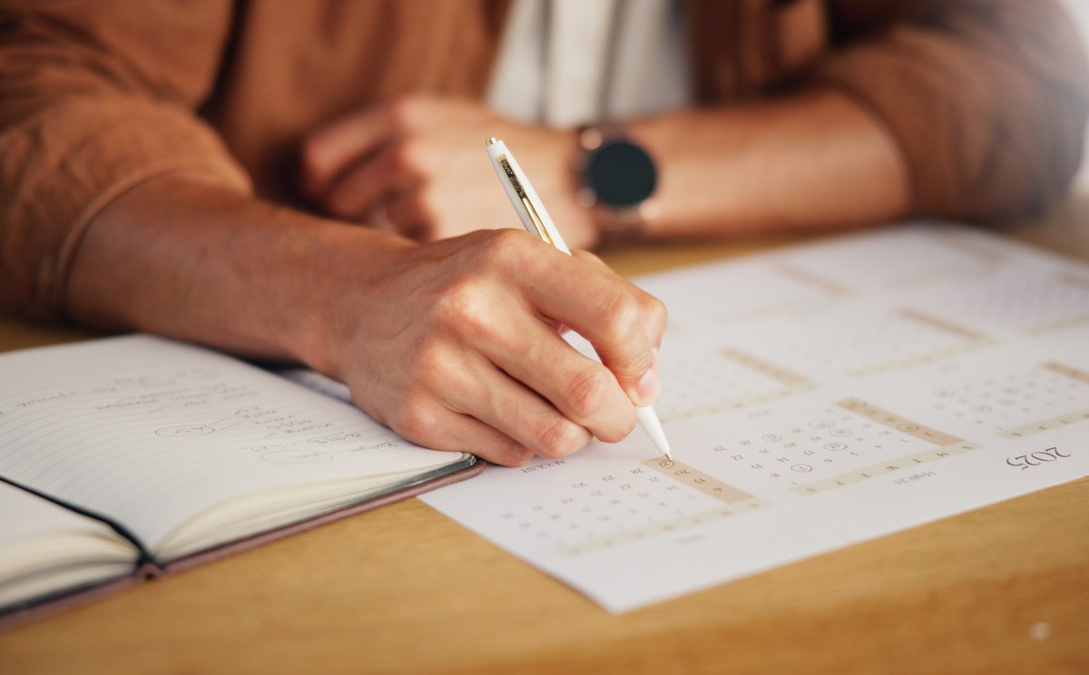 Closeup of a person's hand marking a date on a calendar next to an open planner.