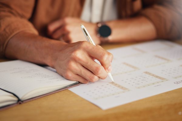 Closeup of a person's hand marking a date on a calendar next to an open planner.