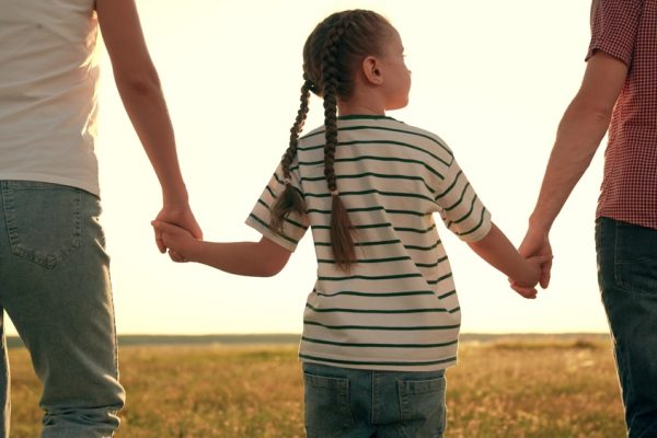 Young girl holding her parents' hands standing in front of a field.