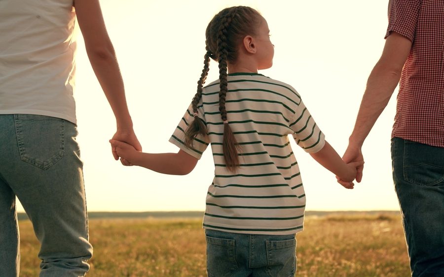 Young girl holding her parents' hands standing in front of a field.
