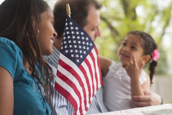 A husband and wife with their daughter sit at a park bench outside with an American flag placed in front of them.