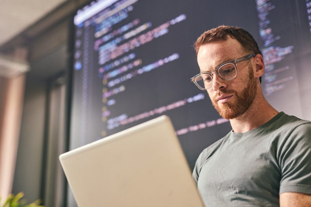 Software developer concentrating on his laptop, with lines of code displayed on a large screen in the background.