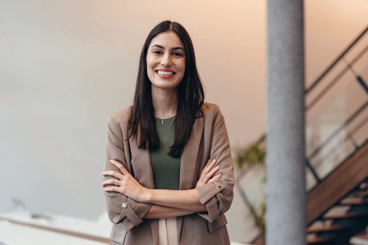 Confident young businesswoman smiling at the camera while standing with arms crossed in a sleek modern office.